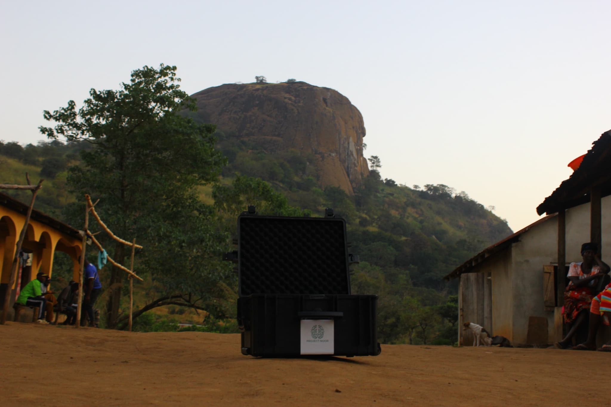 The open transport case stands on a village's red earth square at dusk, houses and a granite inselberg behind it.