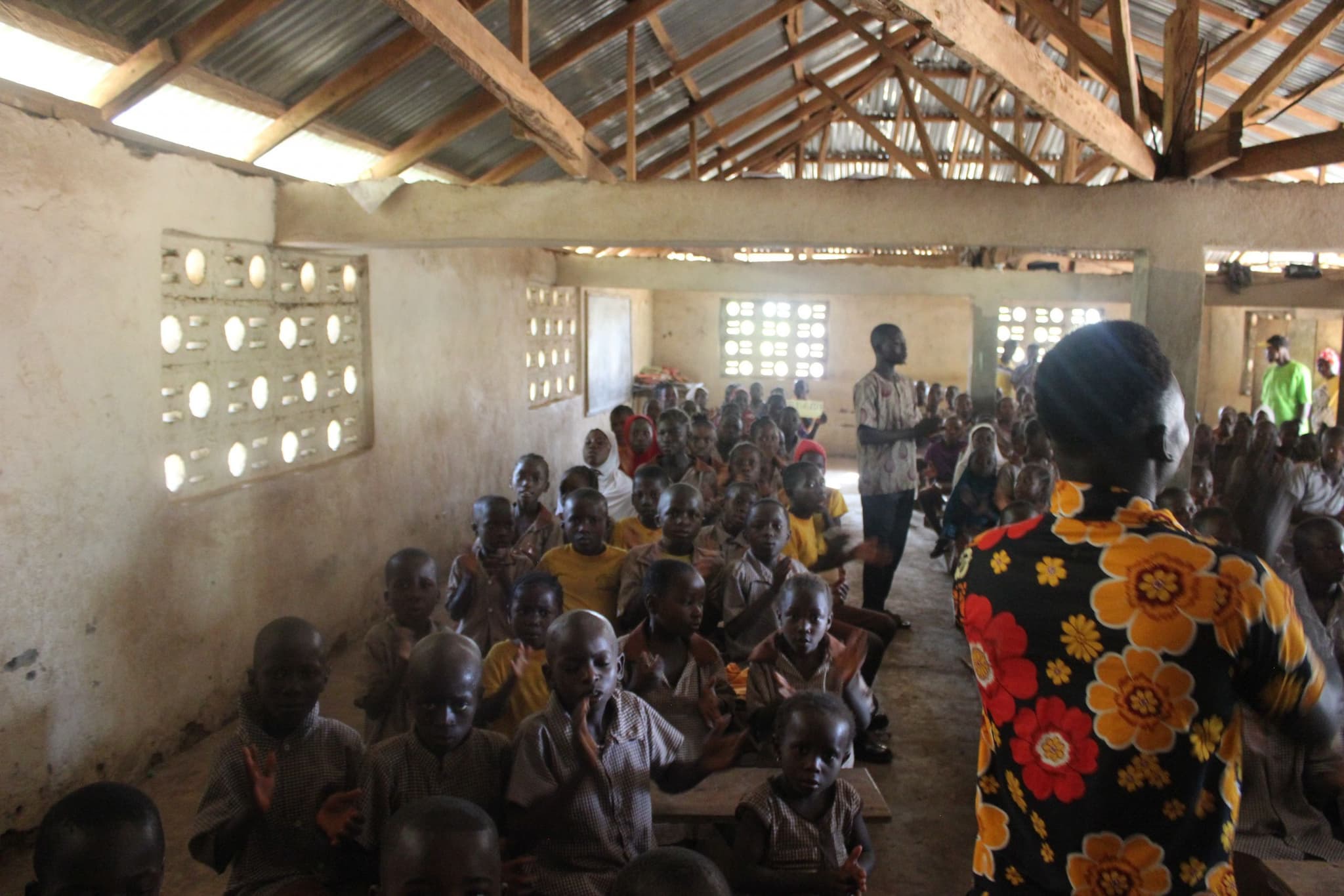 A classroom interior under a corrugated roof, lit only through pierced-block windows, with more children than desks.