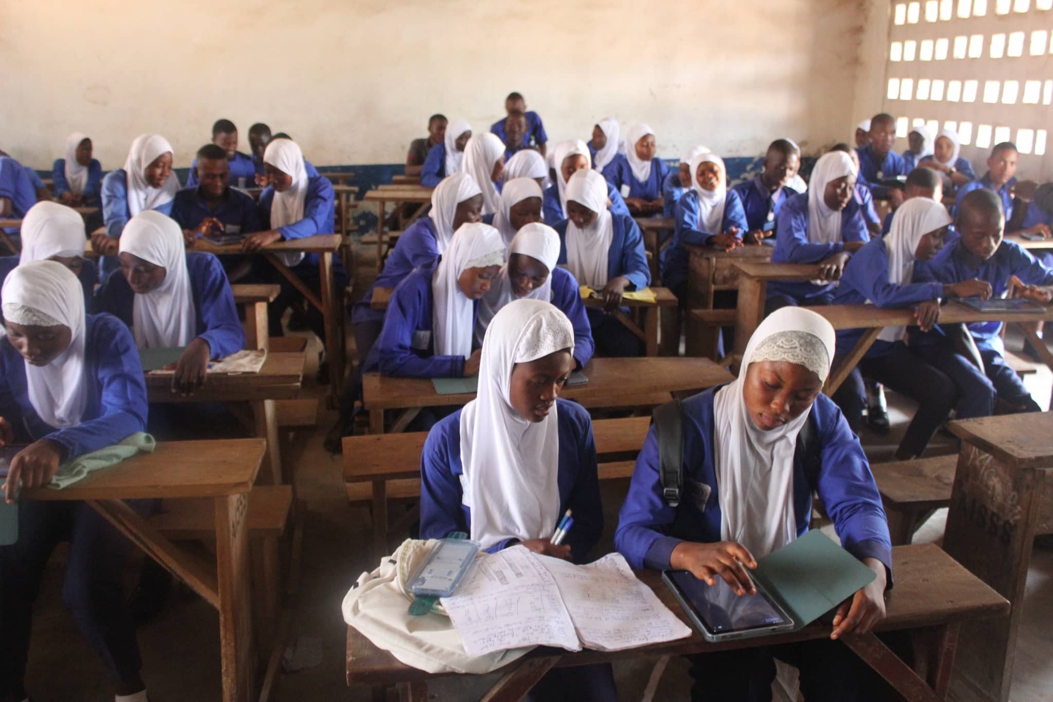 A full classroom: students in blue uniform at wooden benches, several working on tablets beside handwritten exercise books.