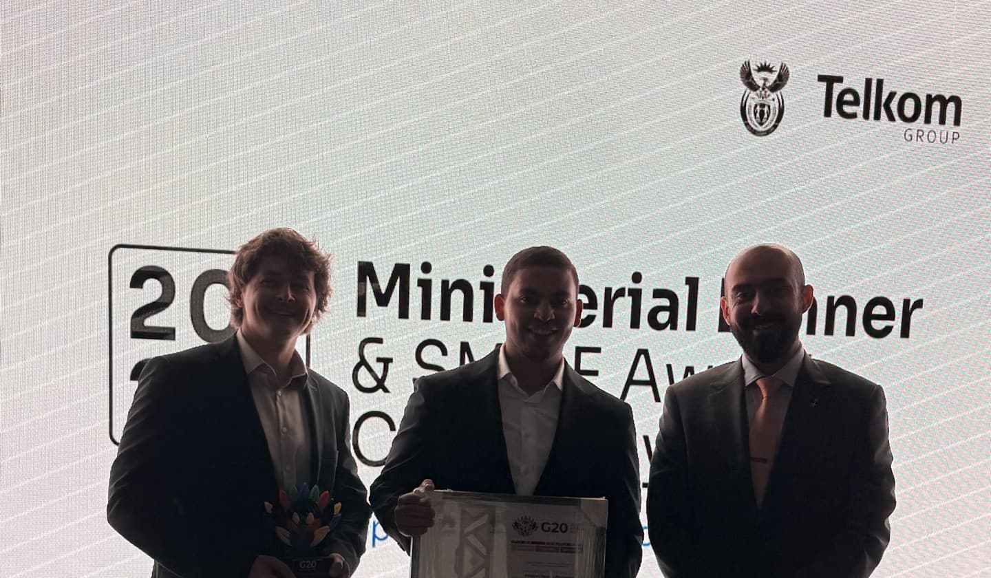Three men in dark suits on stage, the middle one holding the framed certificate; the awards ceremony screen behind them.