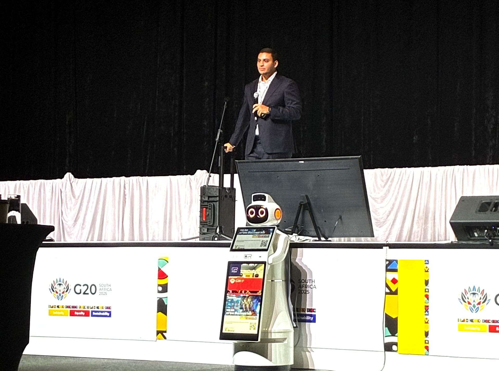 A speaker alone on the G20 stage with a handheld microphone, the stage skirting with its protea motif in front.