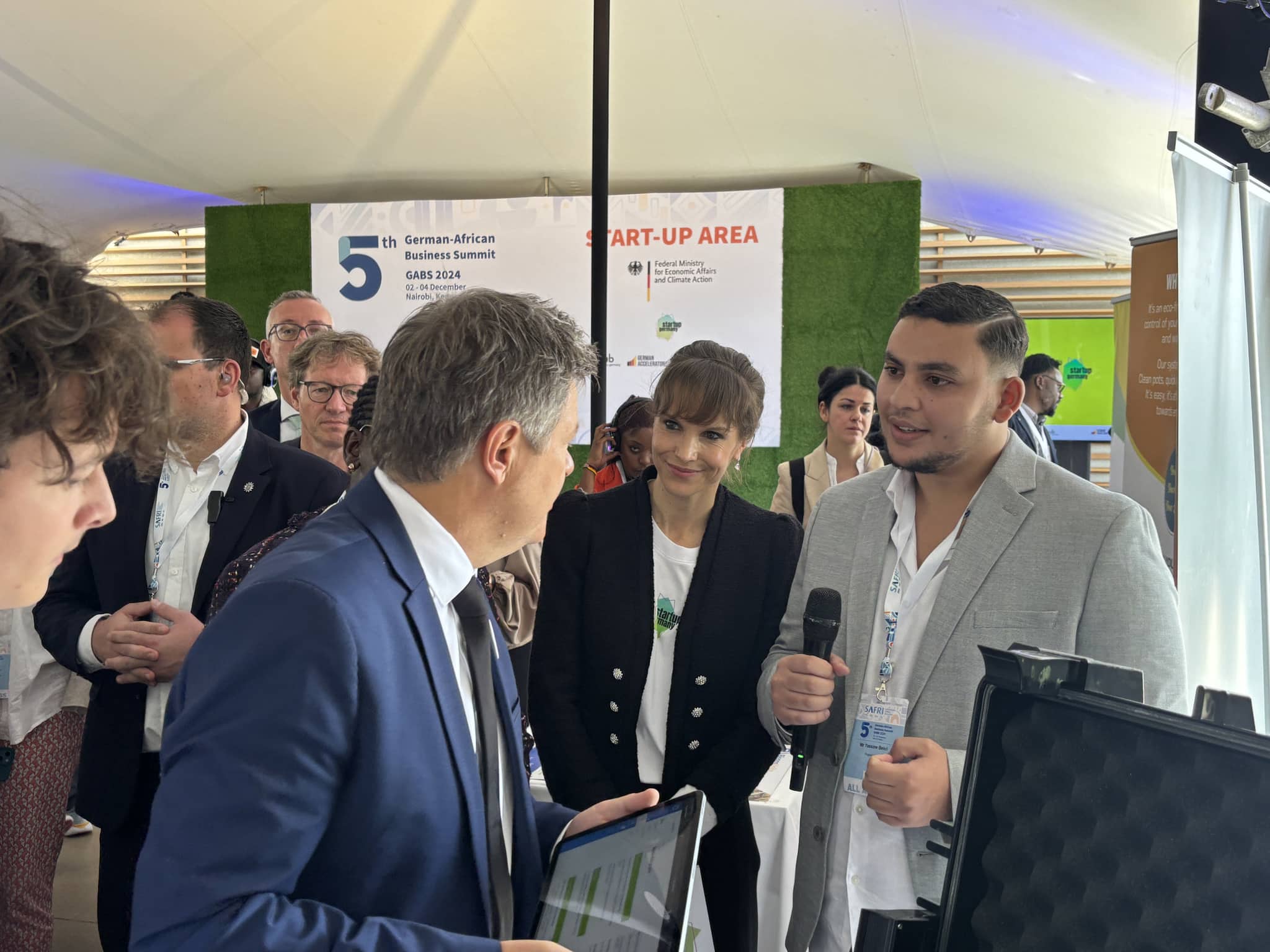 Yassine Bekri, holding a microphone, presents the tablet to the then Vice-Chancellor Robert Habeck at the Project Noor stand; the GABS 2024 banner stands behind them.