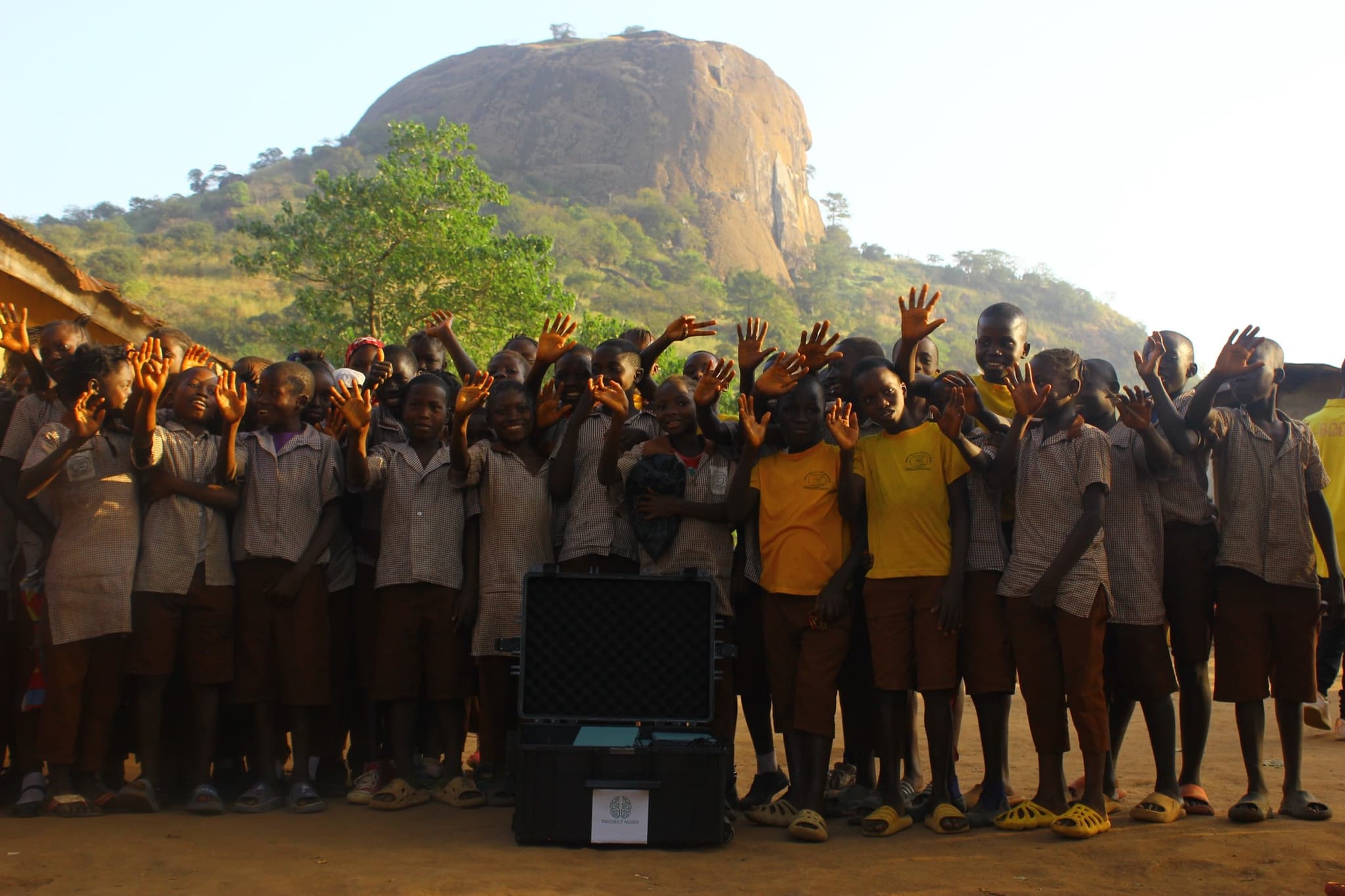 A group of schoolchildren in uniform waving with raised hands; the black Project Noor transport case stands on red earth in front of them, a wooded granite inselberg behind in the evening light.