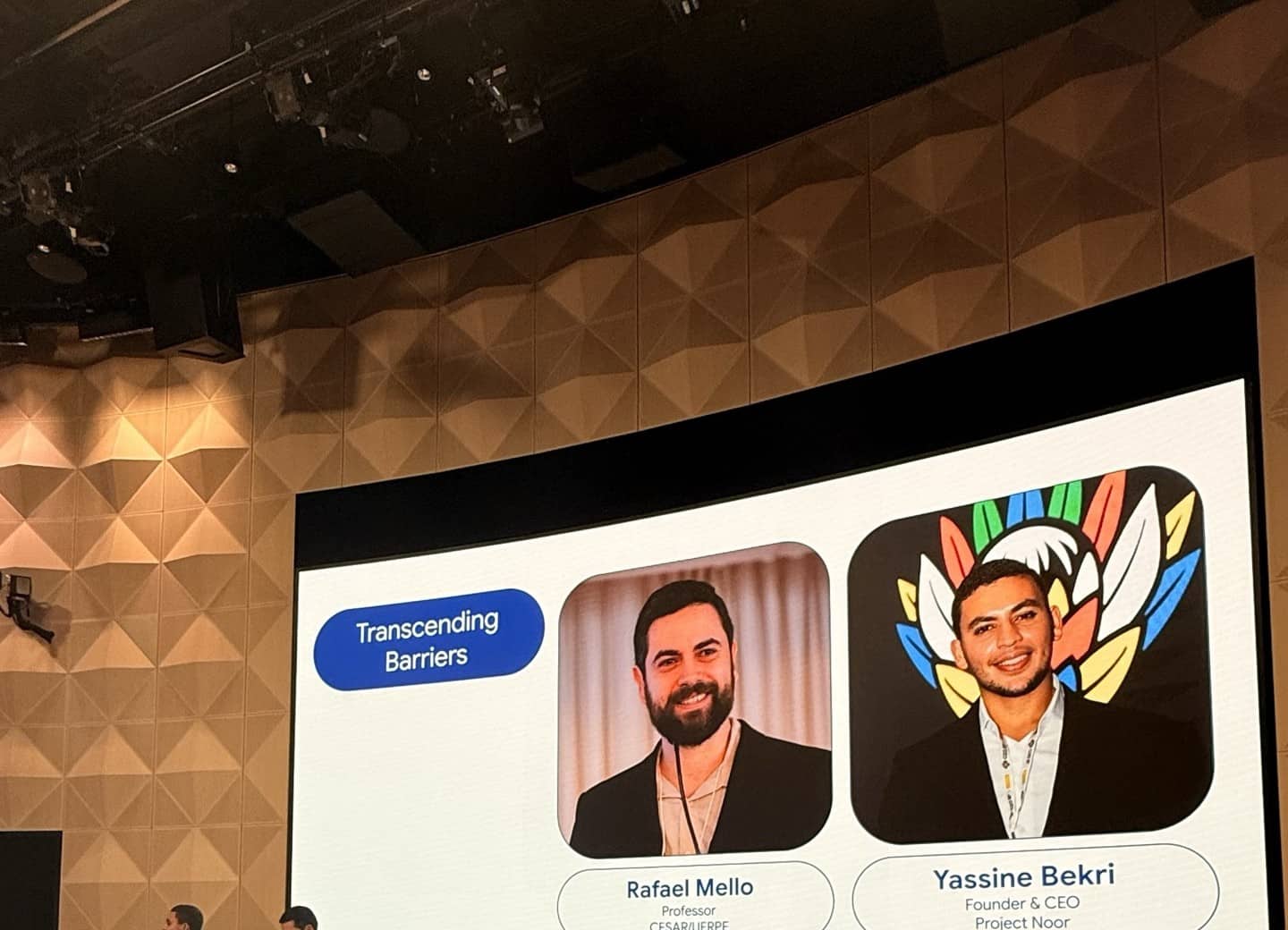 Two speakers stand at a lectern in front of a screen showing the session slide “Transcending Barriers” with two portrait cards.