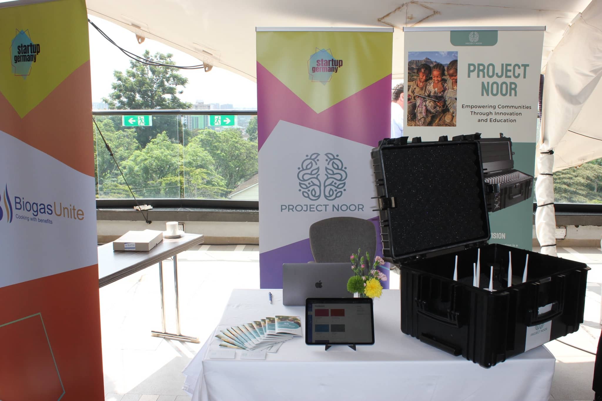 The Project Noor stand: roll-up banners, the open transport case with its charging slots, and a tablet showing the learning platform on the table.