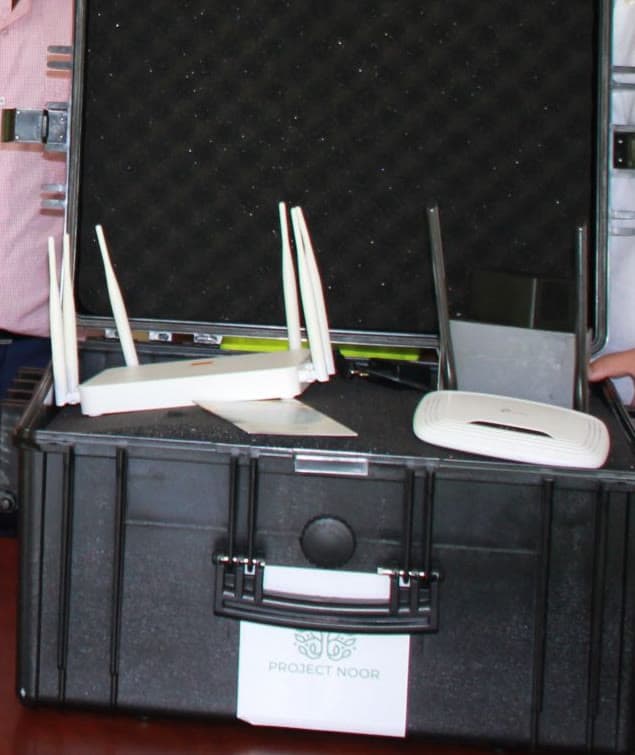 An open black transport case stands on a table; two Wi-Fi routers with antennas and an access point sit in the foam, and a plate carrying the Project Noor mark is fixed to the front. People stand at the left and right edges of the frame.