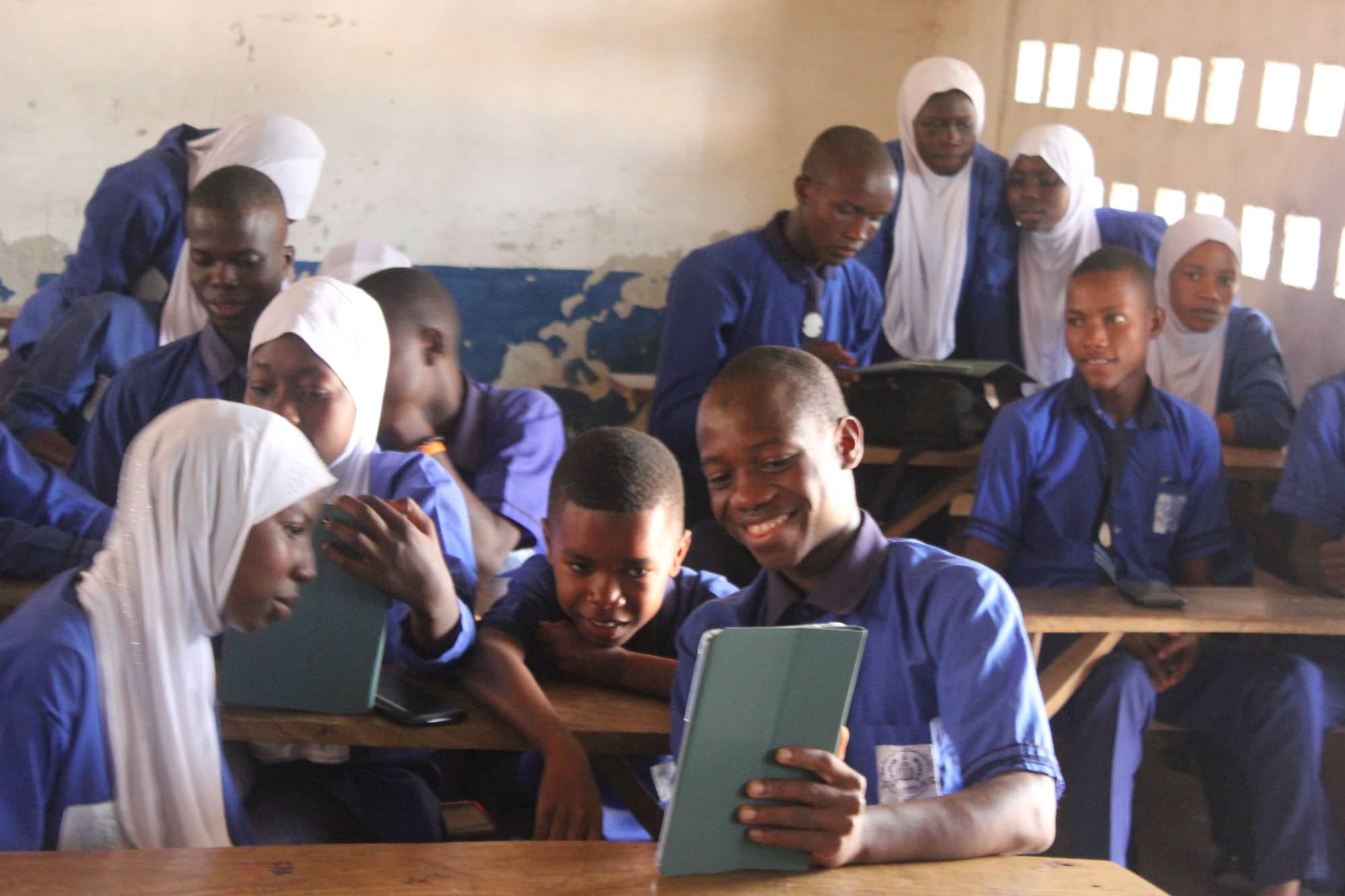A student smiles at the tablet in his hand while classmates in blue uniforms lean in around him.