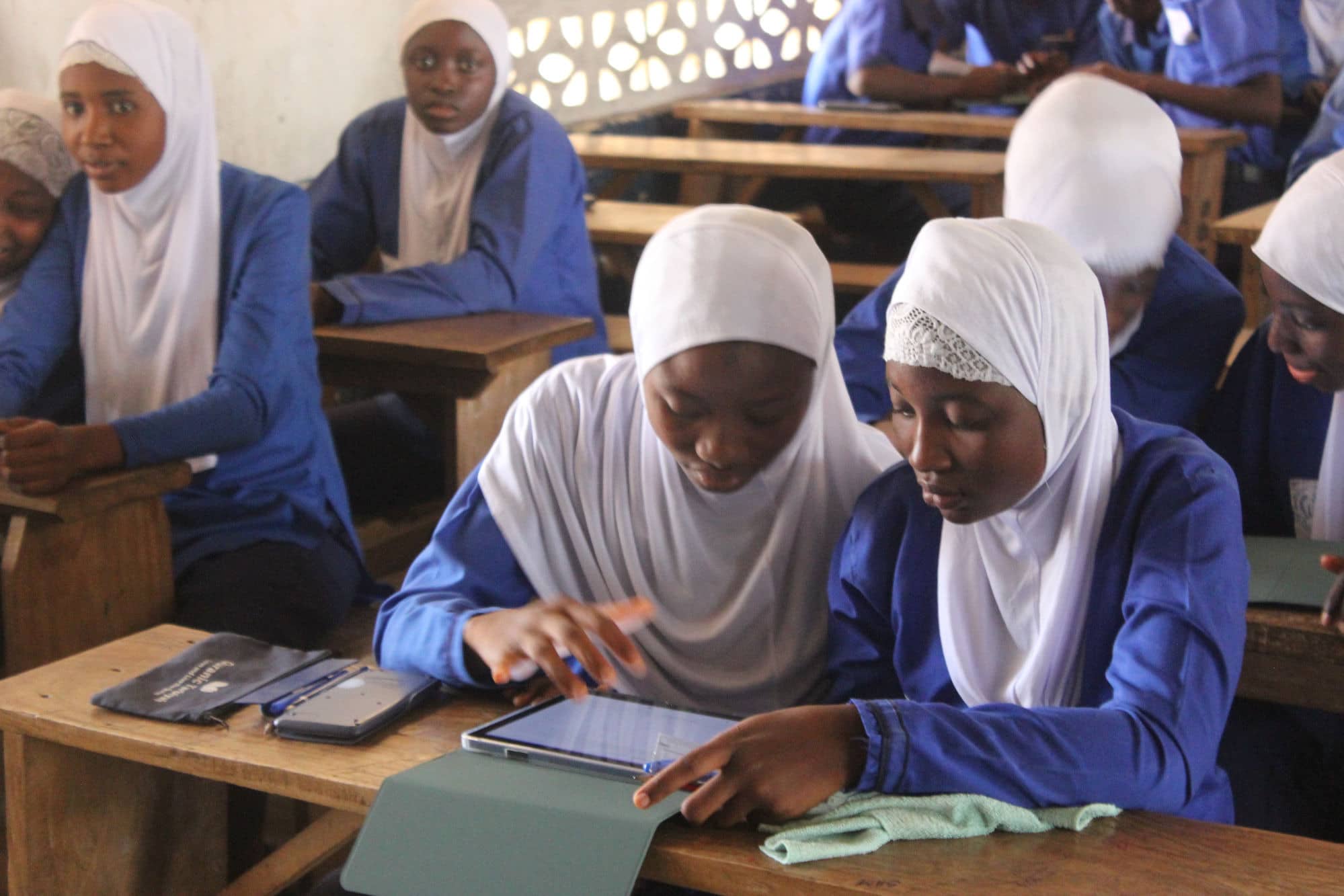 Two students in white headscarves lean over one tablet at a wooden desk, one operating the screen.