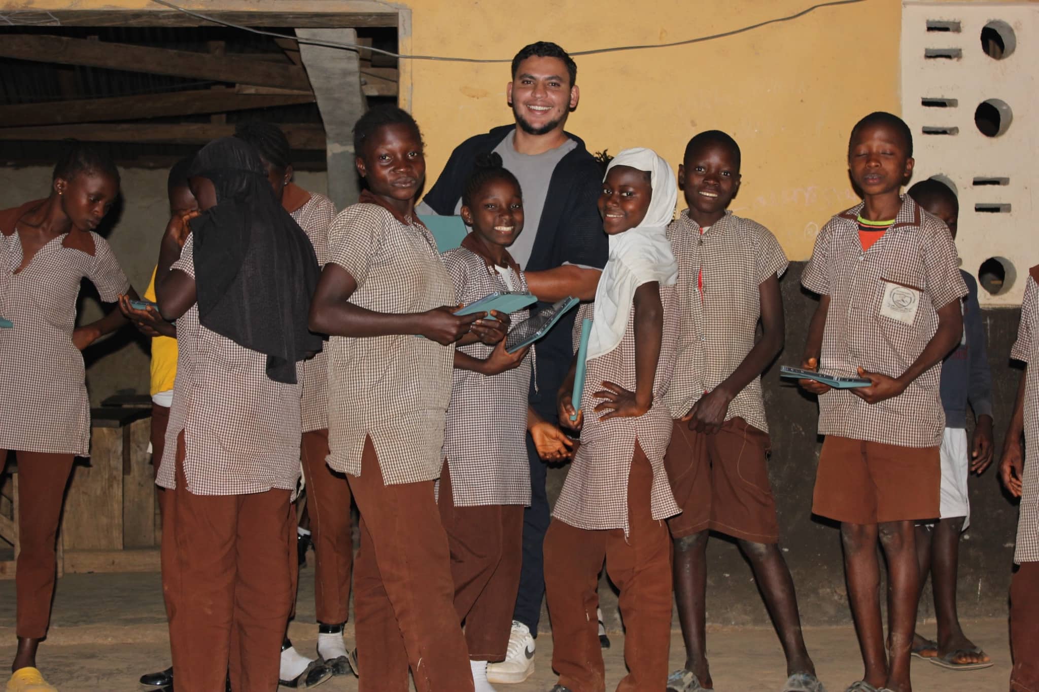A member of the Project Noor team stands among schoolchildren in front of a yellow school building, handing out teal tablets.
